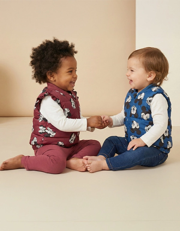 Dwoje maluchów siedzi na jasnym tle, ma na sobie kolorowe kamizelki, trzyma się za ręce i uśmiecha się do siebie.