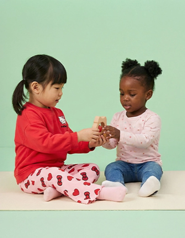 Dwoje małych dzieci siedzi na miękkiej podkładce i radośnie bawi się razem, trzymając kolorową zabawkę na tle w pastelowej zieleni.
