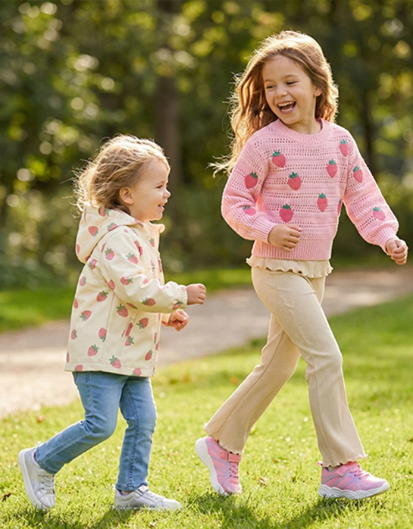 Dwoje dzieci biegnie radośnie po skąpanym w słońcu parku, otoczonym zieloną trawą i drzewami, ubrane w kolorowe swetry i spodnie dresowe.