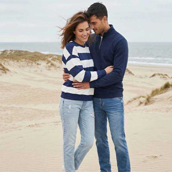 Para w casualowych ubraniach obejmuje się na piaszczystej plaży z wydmami i falami oceanu pod pochmurnym niebem.