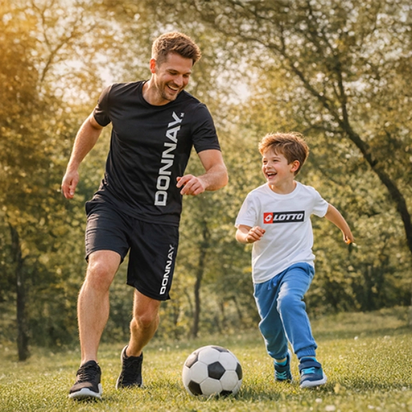Mężczyzna i dziecko radośnie biegają razem po trawiastym boisku, kopiąc piłkę nożną w ciepłym blasku słońca.