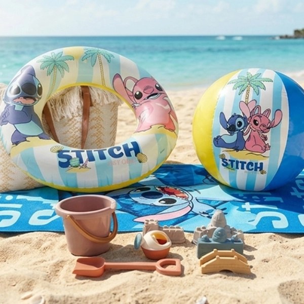 Ręcznik plażowy, koło do pływania, piłka i zabawki do piasku z motywem Lilo i Stitch leżą na piaszczystej plaży przy turkusowym morzu.