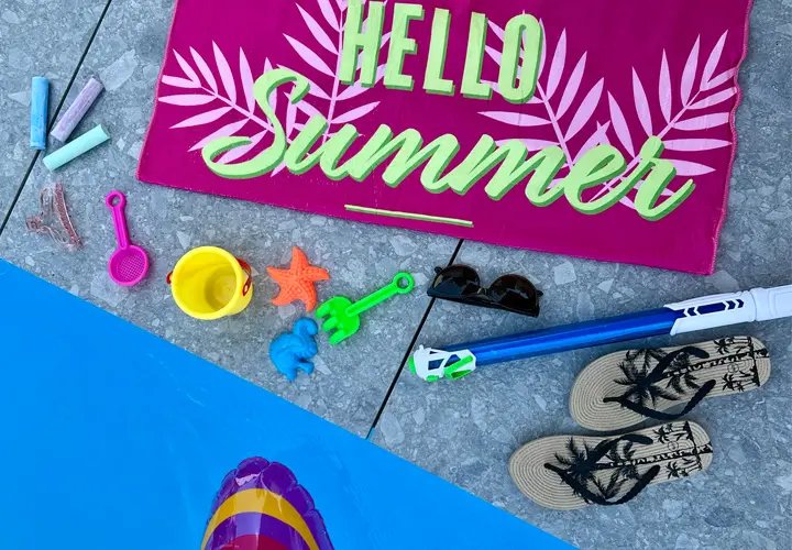 Żywa letnia scena przedstawiająca różowy ręcznik z napisem „Hello Summer”, zabawki plażowe, okulary przeciwsłoneczne i klapki przy basenie.