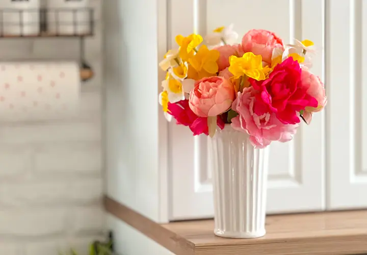 Kolorowe sztuczne kwiaty w białym wazonie, wyeksponowane na drewnianej półce na tle jasnej kuchni.