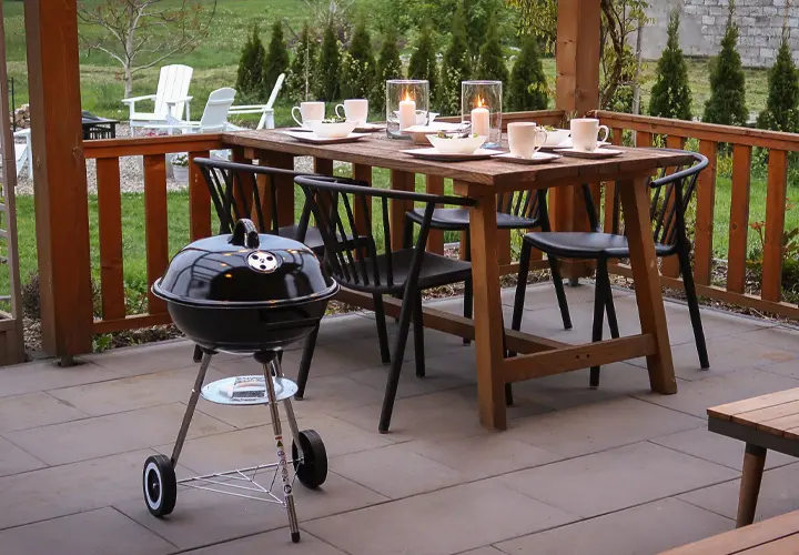 Przytulna jadalnia na świeżym powietrzu z drewnianym stołem ozdobionym świecami, krzesłami i małym czarnym grillem obok – wszystko w otoczeniu zielonego ogrodu.