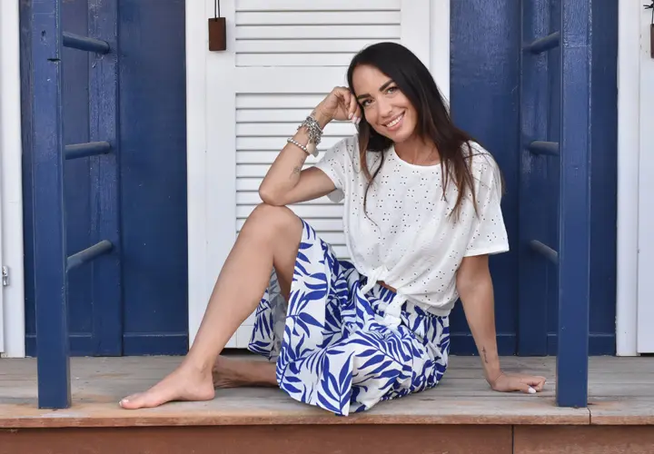 Kobieta siedzi z gracją na werandzie, ubrana w białą bluzkę i niebieską spódnicę w kwiaty, z ręką opartą na brodzie, głęboko zamyślona.