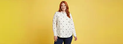 Osoba stoi pewnie przed jasnoczerwonym tłem, ubrana w białą koszulkę z długim rękawem i czarną kamizelkę.