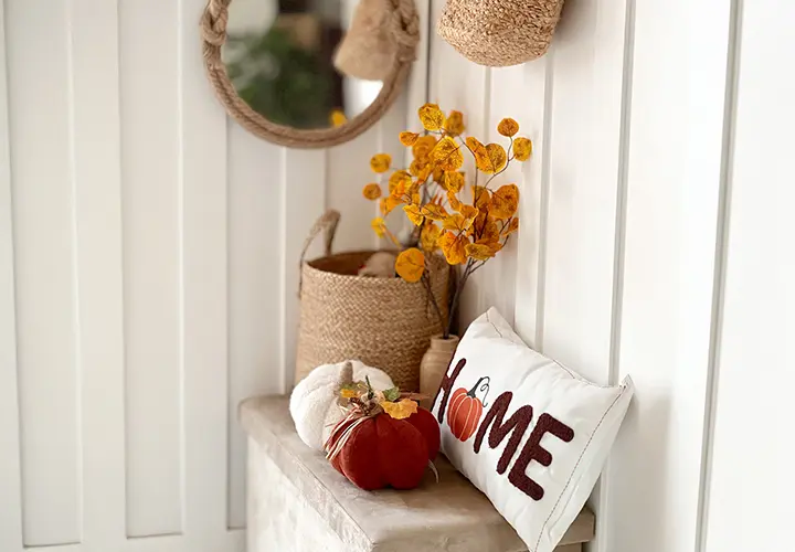 Przytulny wystrój wnętrza z okrągłym lustrem, plecionymi koszami, jesiennymi liśćmi i ozdobną poduszką z napisem „HOME” i akcentem w postaci dyni.