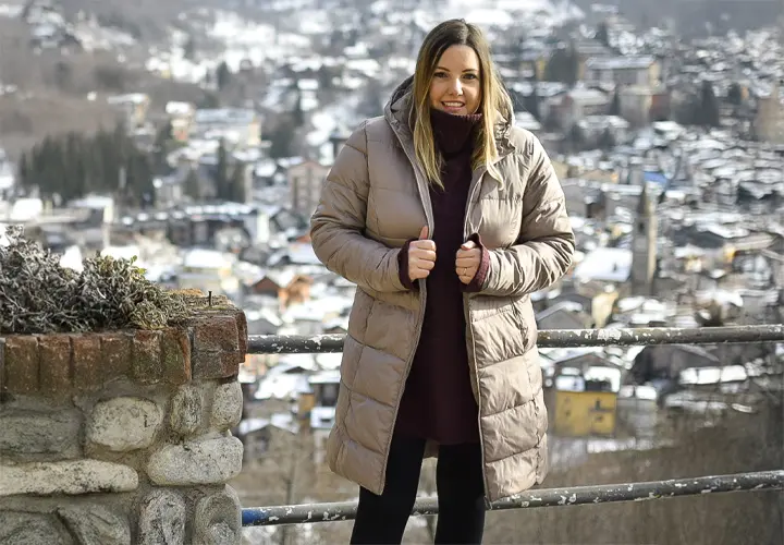 Kobieta w stylowej beżowej kurtce puchowej stoi przy kamiennej balustradzie, spoglądając na ośnieżoną, malowniczą górską wioskę.
