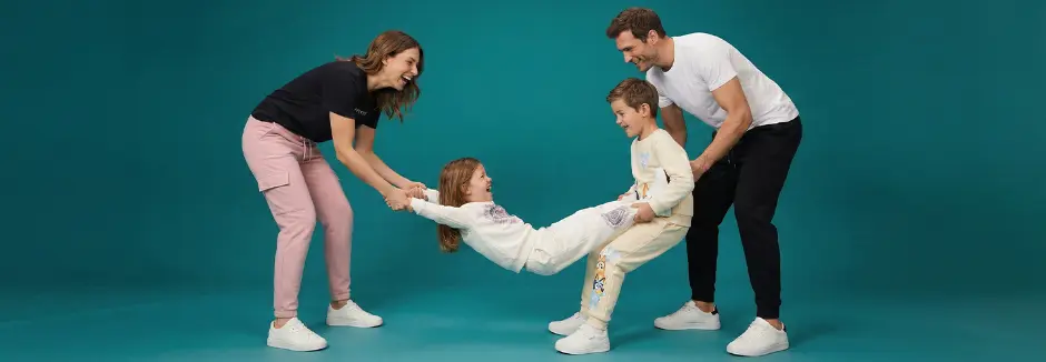 Rodzina bawi się razem, trzymając dziecko między dwojgiem dorosłych; wszyscy są swobodnie ubrani na tle turkusowego tła.