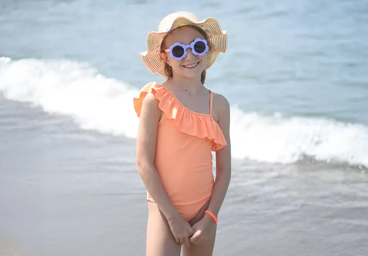Dziecko w koralowym jednoramiennym kostiumie kąpielowym z falbankami stoi na plaży i ma na sobie kapelusz z szerokim rondem. Widoczna jest metka z ceną.