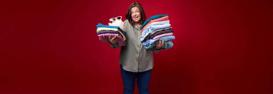 Osoba w szarej koszulce trzyma duże stosy kolorowych, starannie poskładanych ubrań, na których leżą stylowe okulary przeciwsłoneczne i kapelusz.