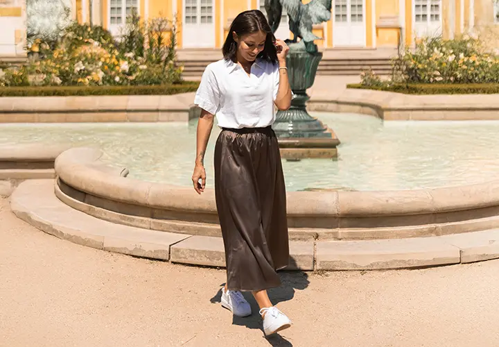 Kobieta w białej koszulce i brązowej spódnicy spaceruje w pobliżu fontanny otoczonej zielenią, w tle żółte budynki.