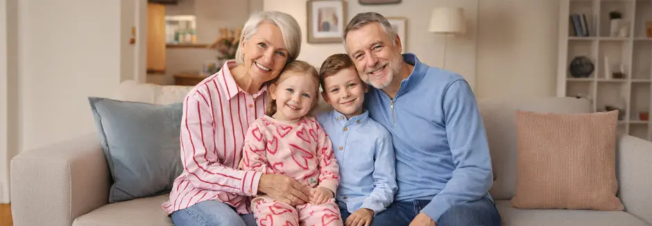 Czteroosobowa rodzina siedzi razem na sofie, uśmiechając się, z miękkimi poduszkami i przytulnym salonem w tle.