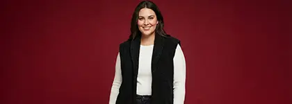 Osoba stoi pewnie przed jasnoczerwonym tłem, ubrana w białą koszulkę z długim rękawem i czarną kamizelkę.