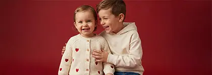 Dwoje dzieci pozuje blisko siebie, jedno w przytulnym swetrze w serduszka, a drugie w jasnej bluzie z kapturem, na czerwonym tle.