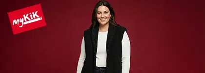 Osoba stoi pewnie przed jasnoczerwonym tłem, ubrana w białą koszulkę z długim rękawem i czarną kamizelkę.