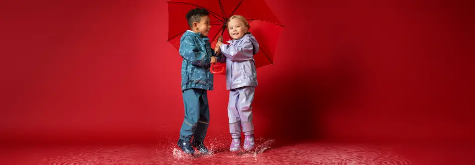 Dwoje dzieci w kolorowych płaszczach przeciwdeszczowych radośnie pluska się w kałużach, dzieląc się jaskrawoczerwonym parasolem.