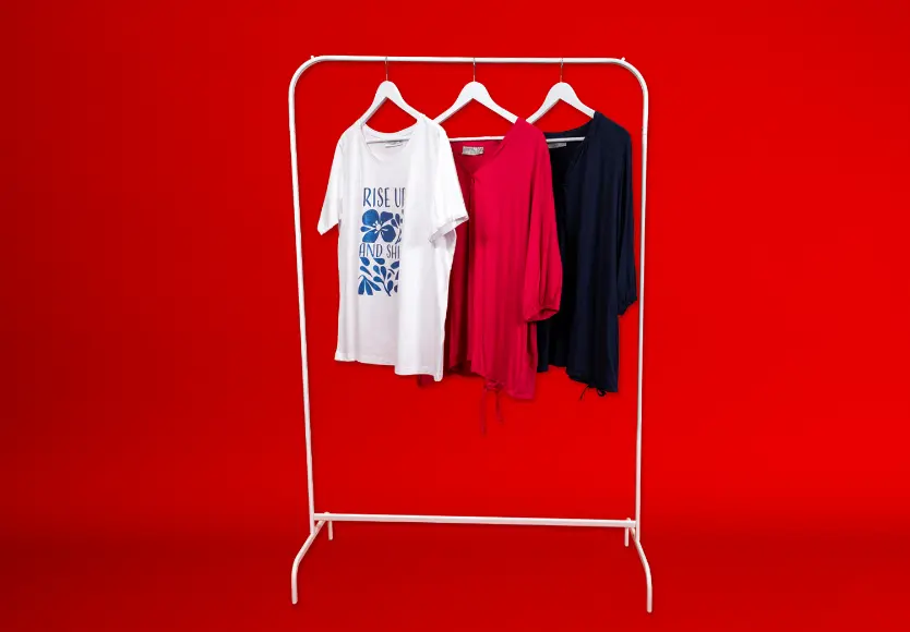 Trzy stylowe topy wiszą na białym wieszaku na tle intensywnie czerwonym: biała koszulka z nadrukiem, różowa bluzka i granatowa koszula.