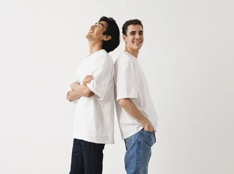 Dwóch młodych mężczyzn w białych koszulkach stoi tyłem do siebie na białym tle i uśmiecha się swobodnie.