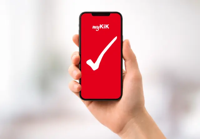 Osoba trzyma smartfon z wyświetloną aplikacją myKiK, mającą czerwone tło z białym znacznikiem wyboru.