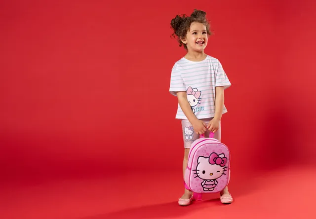 Dziecko stoi przed czerwonym tłem, ubrane w koszulkę w paski z Hello Kitty i szorty, trzymając różowy plecak z Hello Kitty.