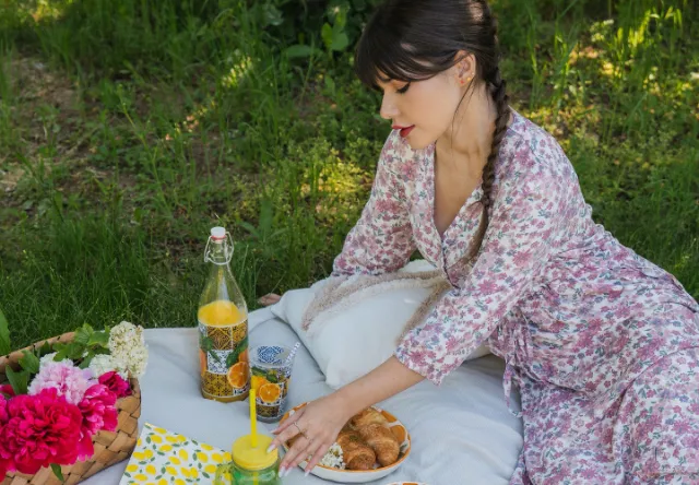 Scena piknikowa przedstawiająca kobietę w sukience w kwiaty, otoczoną bujnymi kwiatami, pomarańczowym napojem, rogalikami i kolorowym kocem na trawie.