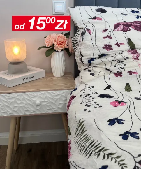 Przytulna scena przy łóżku z zapaloną lampką, wazonem z różowymi różami i wzorzystą kołdrą na tle stonowanej, neutralnej ściany.