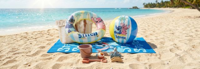 Ręcznik plażowy, koło do pływania, piłka i zabawki do piasku z motywem Lilo i Stitch leżą na piaszczystej plaży przy turkusowym morzu.