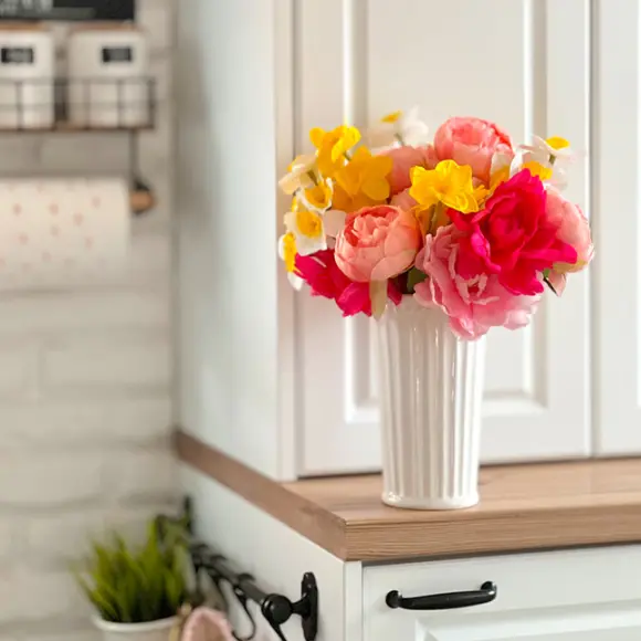 Kolorowe sztuczne kwiaty w białym wazonie, wyeksponowane na drewnianej półce na tle jasnej kuchni.