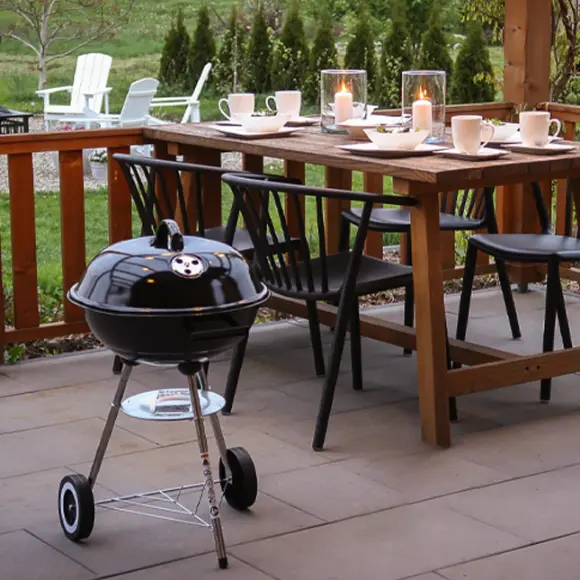 Przytulna jadalnia na świeżym powietrzu z drewnianym stołem ozdobionym świecami, krzesłami i małym czarnym grillem obok – wszystko w otoczeniu zielonego ogrodu.