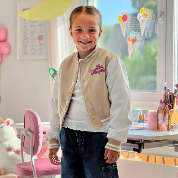 Dziecko w stylowej beżowo-białej kurtce typu varsity stoi w kolorowym, radosnym pokoju pełnym przyborów plastycznych i ozdób.