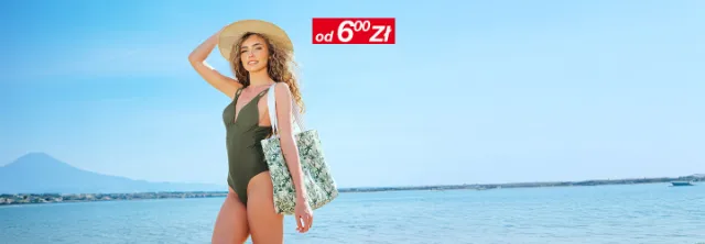 Kobieta w zielonym stroju kąpielowym i kapeluszu z szerokim rondem stoi na plaży, trzymając torbę z tropikalnym wzorem na tle czystego nieba.