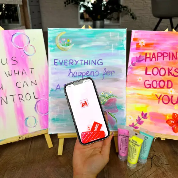 W jednej ręce trzyma smartfon z aplikacją KiK, obok na sztalugach wiszą trzy kolorowe obrazy z motywującymi cytatami.