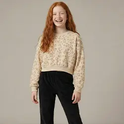 Dziewczynka w beżowym swetrze i czarnych spodniach na jasnym tle.