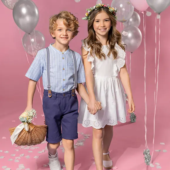 Dwoje dzieci trzyma się za ręce, stojąc na różowym tle. Jedno z nich ma na sobie koszulkę w paski i szorty, a drugie białą sukienkę i kwiecistą koronę.