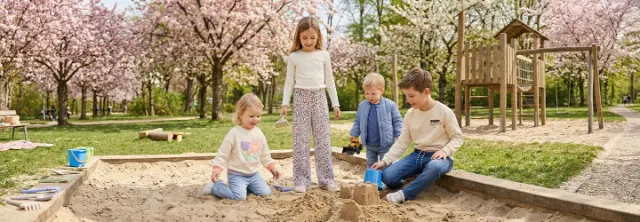 Czworo dzieci bawi się i buduje zamki z piasku w piaskownicy pod kwitnącymi drzewami w parku.