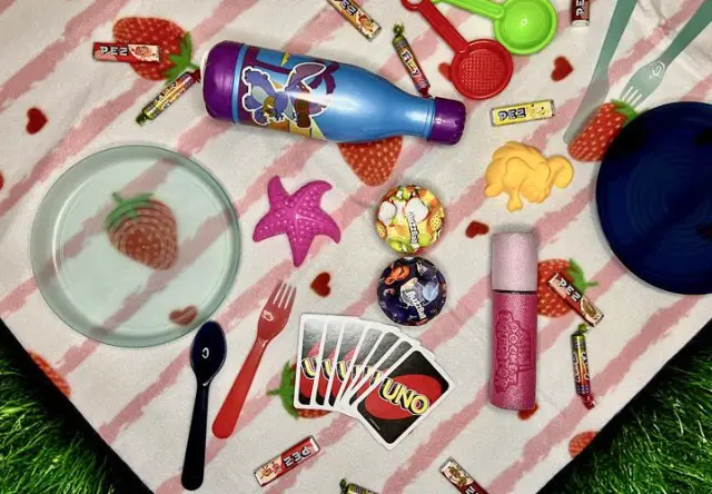 Kolorowy piknik z talerzami, przyborami, słodyczami, zabawkami i butelką napoju na kocu w truskawkowe wzory.