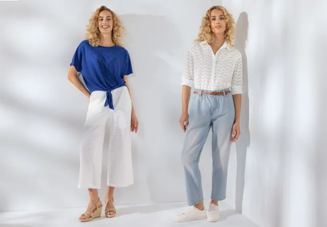 Dwie kobiety prezentują stylowy outfit: jedna w niebieskim topie i białych kulotach, druga w białej bluzce i jasnoniebieskich spodniach na jasnym tle.