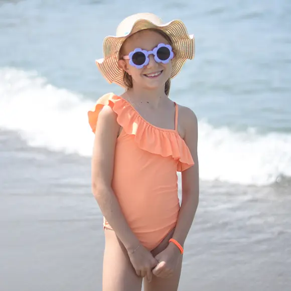 Dziecko w koralowym jednoramiennym kostiumie kąpielowym z falbankami stoi na plaży i ma na sobie kapelusz z szerokim rondem. Widoczna jest metka z ceną.