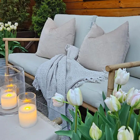 Przytulne miejsce do siedzenia na zewnątrz z miękkimi poduszkami, kocem, świecami i kwitnącymi tulipanami, z wyróżnioną ceną w rogu.