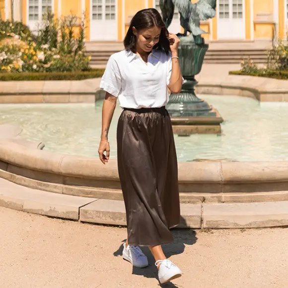 Kobieta w białej koszulce i brązowej spódnicy spaceruje w pobliżu fontanny otoczonej zielenią, w tle żółte budynki.