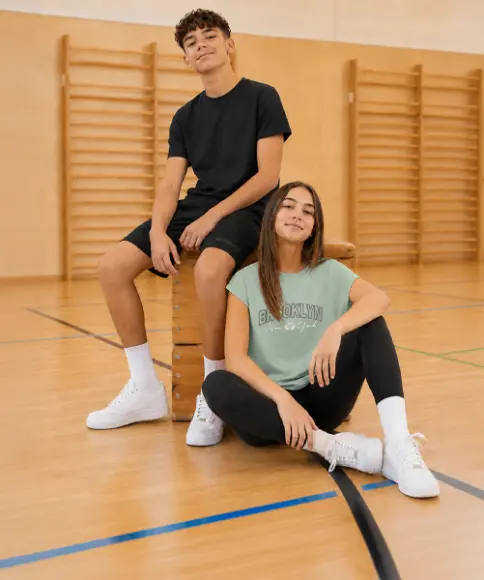 Dwoje nastolatków w wygodnych strojach sportowych siedzi w sali gimnastycznej na i obok skrzyni gimnastycznej. Ubrania doskonale sprawdzą się na lekcjach WF-u.