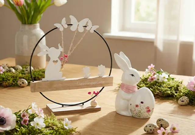 Dekoracyjna wiosenna scenka z figurką królika trzymającego jajko, otoczona motylami, kwiatami i zielenią na drewnianym stole.