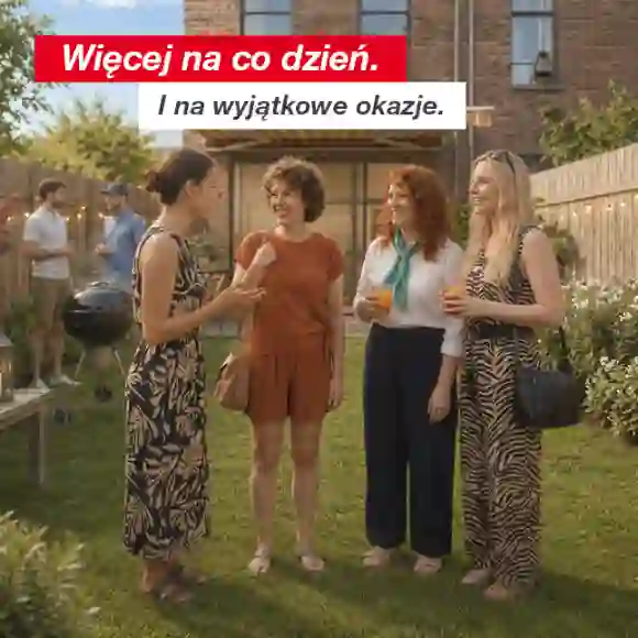Grupa osób rozmawia i pije napoje w ogrodzie na tyłach domu z drewnianym ogrodzeniem i girlandami świetlnymi.