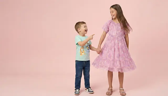 Młody chłopiec w jasnoniebieskiej koszuli i dżinsach wskazuje na dziewczynkę w różowej sukience z falbanami. Oboje trzymają się za ręce na tle delikatnego różowego tła. Młody chłopiec w jasnoniebieskiej koszuli i dżinsach wskazuje na dziewczynkę w różowej sukience z falbanami. Oboje trzymają się za ręce na tle delikatnego różowego tła.
