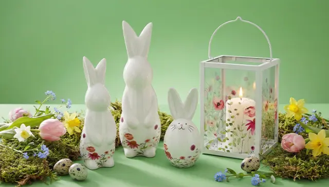Trzy ceramiczne króliczki, wzorzyste jajko i latarnia ze świeczką, otoczone kolorowymi kwiatami i pisankami na zielonym tle. Trzy ceramiczne króliczki, wzorzyste jajko i latarnia ze świeczką, otoczone kolorowymi kwiatami i pisankami na zielonym tle.