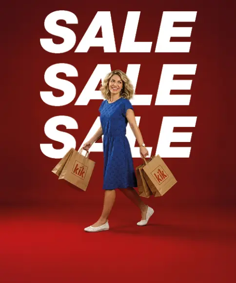Kobieta w niebieskiej sukience niesie kilka toreb zakupowych z logo KiK i idzie na tle dużego białego napisu „SALE” na czerwonym tle.