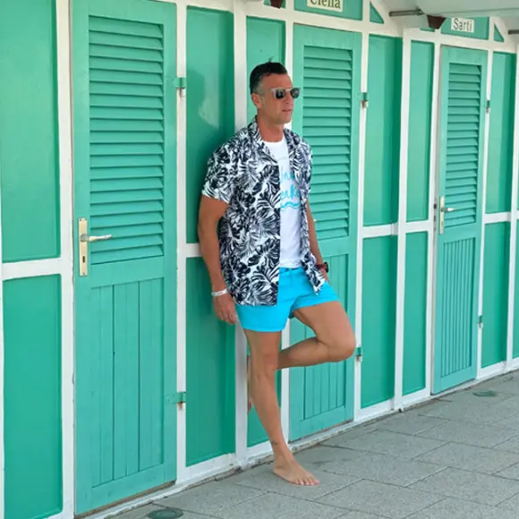 Mężczyzna w tropikalnej koszuli i niebieskich szortach swobodnie opiera się o zielone domki plażowe i cieszy się słonecznym dniem.