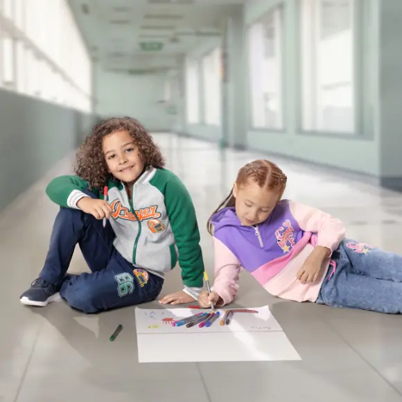 Dwoje dzieci siedzi na podłodze wyłożonej kafelkami, rysując kolorowymi markerami na dużym arkuszu papieru.